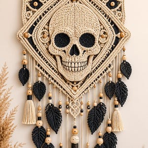 May include: A macrame wall hanging featuring a central crocheted skull in beige yarn with black eye and nose sockets. The skull is framed by a diamond-shaped macrame pattern with wooden beads, black yarn leaves, and beige tassels hanging below.