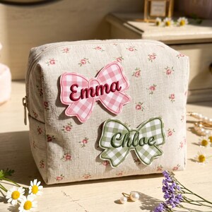May include: A beige fabric cosmetic bag with a small pink floral pattern. The bag features two bow-shaped patches: a pink gingham bow with the embroidered name Emma and a sage green gingham bow with the embroidered name Chloe in dark green.