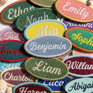 May include: A collection of oval-shaped embroidered patches featuring various names in white or colored cursive script. Each patch has a distinct border color, including orange, green, yellow, light blue, and purple, with names like Ethan, Emily, Noah, Mia, Benjamin, and Liam.