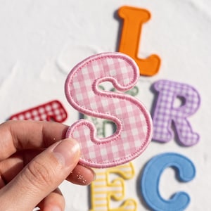 May include: A pink and white gingham fabric letter S patch with a stitched border. Other fabric letter patches in orange, purple, red, blue, and yellow with various patterns are visible in the background, including a letter L, R, and C.