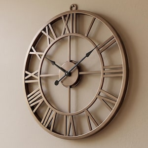May include: A large, circular wall clock with a metallic bronze finish. The clock features an open-frame design with prominent Roman numerals, thin black metal hands, and a central gear-like detail. A small circular loop is at the top for hanging.