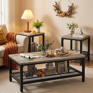 May include: A rectangular coffee table and two matching square end tables featuring weathered gray wood-grain surfaces and black metal frames. Each table includes a lower mesh storage shelf holding books, a calendar, and decor.