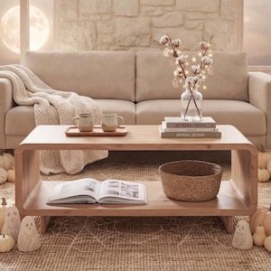 May include: A light wood coffee table with a lower shelf, holding two mugs on a tray, a stack of books, a glass vase with cotton stems and fairy lights, and a woven basket. An open book rests on the lower shelf, surrounded by ghost figurines.
