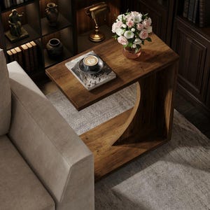 May include: A C-shaped wooden side table with a dark brown finish and a curved support base. The tabletop holds a bouquet of pink and white roses in a copper-toned vase, a ceramic cup of coffee on a saucer, and a book with a black and white cover.