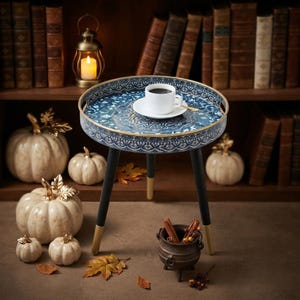 May include: A round side table with a blue and white patterned tray top and three black legs with gold-toned tips. A white ceramic cup filled with dark coffee sits on a matching saucer in the center of the table, which features a raised metal rim.