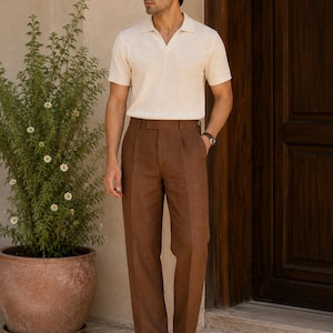 Könnte beinhalten: Ein cremefarbenes Kurzarm-Poloshirt mit gerippter Textur und V-Ausschnitt, kombiniert mit einer braunen Hose mit Faltenfront und geradem Bein. Das Outfit wird durch braune Ledersandalen und eine dunkle Sonnenbrille vervollständigt.