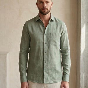 May include: A long-sleeved button-down shirt in a muted sage green color. The shirt features a classic collar, a single chest pocket on the left side, and a row of matching buttons down the front. The fabric has a visible, slightly textured weave.