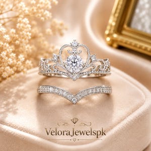 May include: A silver-toned ring set consisting of a crown-shaped band featuring a central round-cut stone and intricate filigree detailing, paired with a matching V-shaped chevron band encrusted with small clear stones along the entire surface.