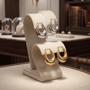 May include: Two pairs of hoop earrings displayed on a beige stand. The top pair is silver-toned with a smooth, polished finish, while the bottom pair is gold-toned with a matching smooth, reflective surface. Both sets feature a thick, teardrop shape.