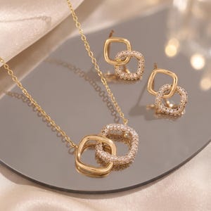 May include: A gold-toned necklace and matching stud earrings set. Each piece features two interlocking square-shaped links, with one link having a smooth polished finish and the other encrusted with small, clear, sparkling stones.