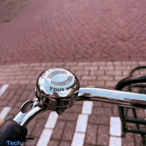 Può includere: Un campanello per bicicletta in cromo lucido montato su un manubrio argentato. La parte superiore del campanello presenta il testo nero "PUT A BELL ON IT TOGETHER YOUR DESIGN". Il campanello ha una forma a cupola arrotondata.