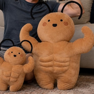 Puede incluir: Dos peluches de color tostado con forma de figuras musculosas con abdominales definidos, cabezas redondas y dos antenas negras. Cada peluche presenta una cara sencilla con dos pequeños ojos negros y mejillas rosadas, imitando una pose.