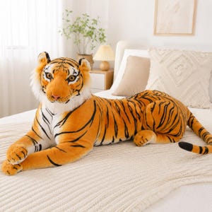 Könnte beinhalten: Ein großes Plüsch-Tiger-Spielzeug mit orangefarbenem Fell mit schwarzen Streifen, einem weißen Bauch und weißem Fell um das Gesicht. Der Tiger liegt in einer liegenden Position mit nach vorne ausgestreckten Pfoten und einem langen Schwanz.