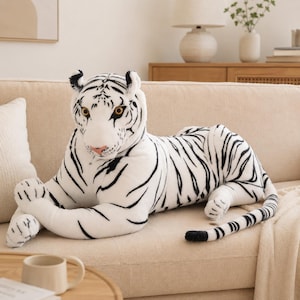 Può includere: Un grande peluche a forma di tigre bianca con strisce nere, occhi color ambra e naso rosa. L'animale di peluche è raffigurato in posizione sdraiata con le zampe anteriori ripiegate verso l'interno e una lunga coda distesa sul divano.