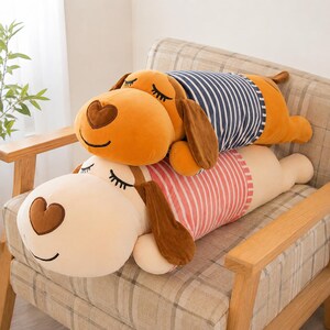 May include: Two long, plush dog-shaped pillows stacked on top of each other. The top dog is orange with a dark blue and white striped shirt. The bottom dog is cream-colored with a pink and white striped shirt. Both have heart-shaped noses.