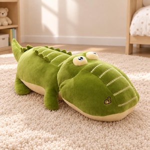 Puede incluir: Un cocodrilo de peluche con un cuerpo de color verde vibrante y una parte inferior de color beige claro. El juguete tiene ojos grandes de estilo caricatura, una serie de crestas suaves en la espalda y un pequeño logotipo bordado en el hocico.