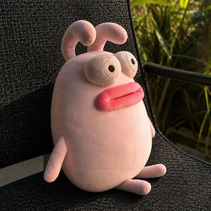 Pode incluir: Um brinquedo de pelúcia rosa claro com corpo arredondado, duas pequenas antenas no topo, olhos brancos grandes com pupilas pretas e lábios grossos e rosa brilhante. O personagem macio tem dois braços e duas pernas pequenas no corpo.