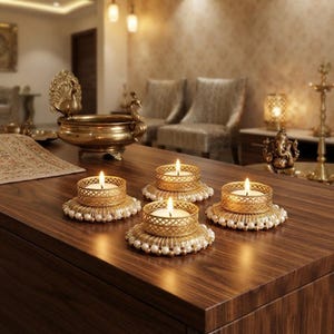Può includere: Tre portacandele in metallo dorato con intricati motivi a reticolo, ciascuno poggiato su una base circolare decorata con un anello di piccole perle bianche. Ogni supporto contiene una candelina bianca accesa che emana luce calda.