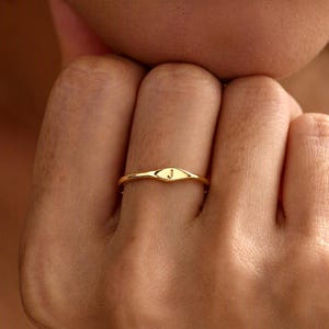 Könnte beinhalten: Ein dünner Goldring mit einer kleinen, flachen, ovalen Oberfläche, in die der Großbuchstabe J eingraviert ist. Das Band ist glatt und poliert, mit einem minimalistischen Design, das mittig auf dem Finger sitzt und ein helles, reflektierendes Finish aufweist.