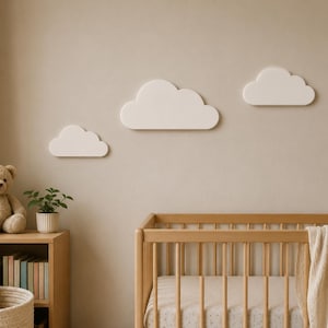 May include: Three white cloud-shaped wall decorations mounted on a beige wall above a light wood cot. The cot features a light-coloured blanket draped over the side, while a teddy bear and a small potted plant sit on a nearby wooden shelf.