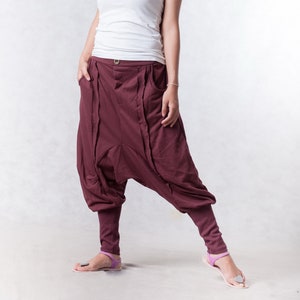 Może przedstawiać: Kobieta w białej koszulce i maronowych spodniach typu harem. Stoi z lekko rozstawionymi nogami i opuszczonymi ramionami. Patrzy w górę i w prawo.