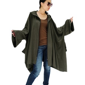 Könnte beinhalten: Eine Frau trägt eine lange, dunkelgrüne Kapuzenjacke mit Reißverschluss. Die Jacke hat eine lockere Passform und lange Ärmel. Sie trägt auch blaue Jeans und schwarze Stiefeletten mit Absatz.
