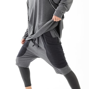 Może przedstawiać: Szara bluza z kapturem z detalem zamka błyskawicznego na ramieniu i szare i czarne spodnie dresowe z dużą kieszenią z boku. Model ma na sobie białe buty sportowe.