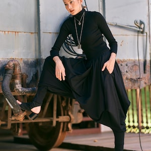 May include: A woman wearing a black turtleneck, long necklace, and black harem pants with stirrup leggings. The outfit is paired with black sandals. The background features a weathered metal structure.