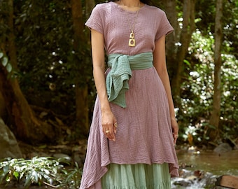 Könnte beinhalten: Eine Frau trägt ein langes, mehrschichtiges Kleid mit einem grünen Gürtel. Das Kleid ist helllila und hat einen Rüschen-Saum. Die Frau steht in einer Waldlandschaft.