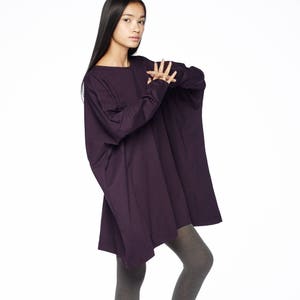 May include: A woman wearing a long, loose-fitting, dark purple tunic top with long sleeves and grey leggings. She is wearing black heeled sandals with ankle straps. The top is styled with a slightly asymmetrical hemline.