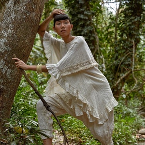 May include: A woman wearing a white, ruffled, crocheted top and white pants stands in a stream in a forest. She is holding a branch with one hand and has her other hand on her head.
