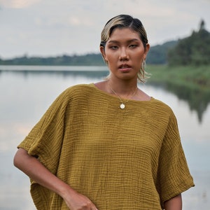 Peut inclure: Une femme portant un haut ample à manches courtes de couleur moutarde avec un tissu texturé. Elle se tient devant une étendue d'eau et porte une jupe marron.