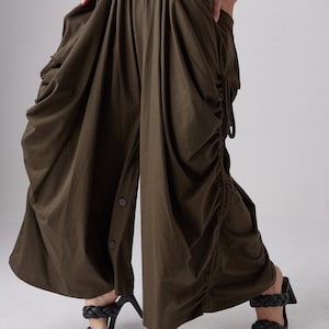 May include: A pair of olive green wide-leg pants with a drawstring waist and a button-up front. The pants are gathered and have a loose, flowing fit.