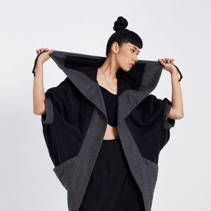 May include: A black and gray hooded cardigan with a long, flowing silhouette. The cardigan has a large hood and a relaxed fit. The model is wearing the cardigan with a black top and black pants.