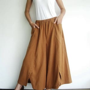 May include: A woman is wearing a white tank top and brown wide-leg pants with a loose, flowing fit. The pants have a wide waistband and pockets on the sides.