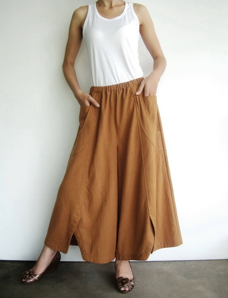 May include: A woman is wearing a white tank top and brown wide-leg pants with a loose, flowing fit. The pants have a wide waistband and pockets on the sides.