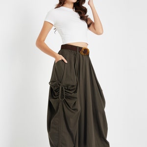 Puede incluir: Una mujer con un top blanco corto y una falda verde oscuro con pliegues y un cintur&oacute;n marr&oacute;n. Lleva zapatos de tac&oacute;n marrones.