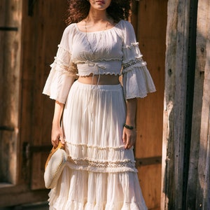 Puede incluir: Un top blanco de crochet con un dobladillo con volantes y mangas largas, combinado con una falda maxi blanca a juego con m&uacute;ltiples niveles de volantes. La falda tiene un borde de encaje y el top tiene un lazo en la cintura.