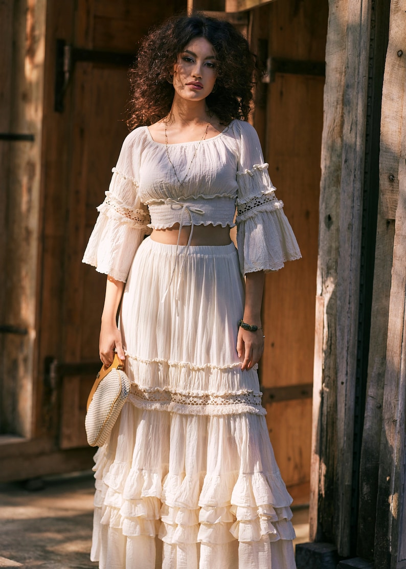 Puede incluir: Un top blanco de crochet con un dobladillo con volantes y mangas largas, combinado con una falda maxi blanca a juego con m&uacute;ltiples niveles de volantes. La falda tiene un borde de encaje y el top tiene un lazo en la cintura.