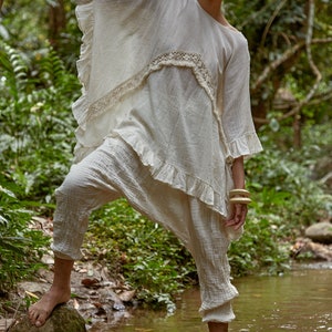 May include: A woman wearing a white, ruffled, crocheted top and white pants stands in a stream. She is holding onto a tree branch with her left hand and has a wooden bracelet on her right wrist.