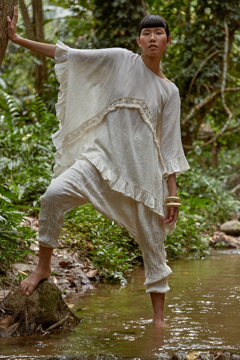 May include: A woman wearing a white, ruffled, crocheted top and white pants stands in a stream. She is holding onto a tree branch with her left hand and has a wooden bracelet on her right wrist.