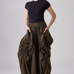 May include: A woman is wearing a black short-sleeved shirt and a long, dark green skirt with a gathered design. The skirt has a drawstring detail on the sides. She is wearing black heeled sandals.