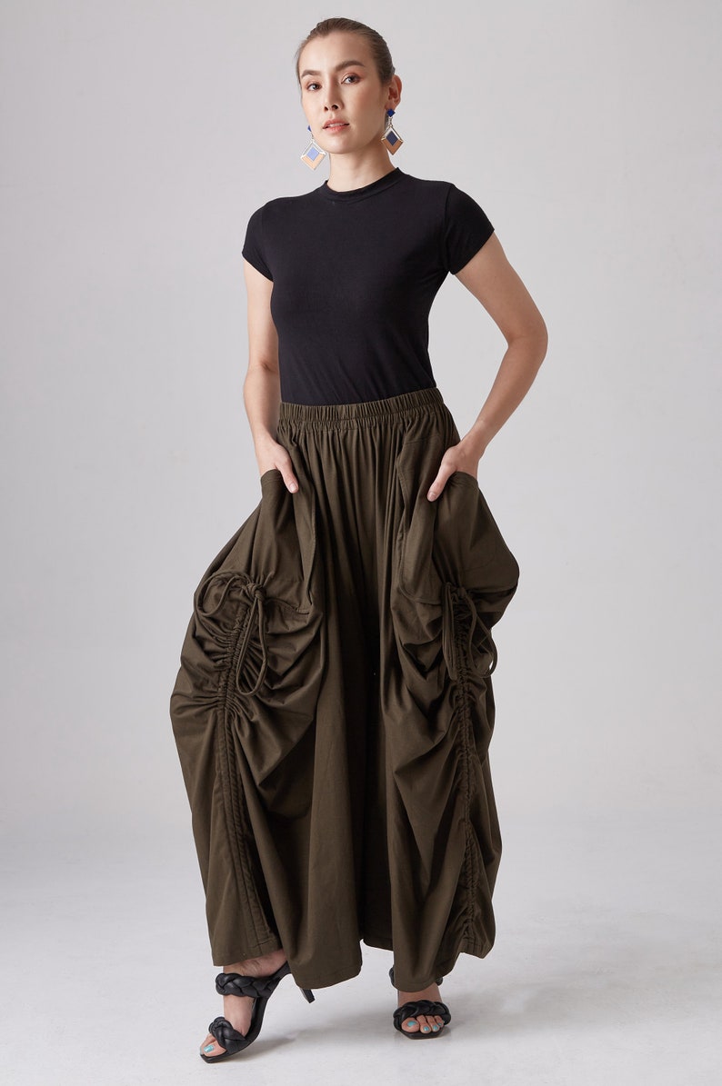 May include: A woman is wearing a black short-sleeved shirt and a long, dark green skirt with a gathered design. The skirt has a drawstring detail on the sides. She is wearing black heeled sandals.