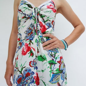 May include: White floral print sundress with spaghetti straps and a lace trim. The dress features a tie detail at the bust and a tiered skirt. The floral pattern includes red, blue, and green accents. A bracelet with blue and white beads is worn on the arm.