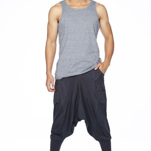 May include: A man wearing a gray tank top and dark blue pants with a unique design. He is standing with his hands in his pockets and looking to the side.