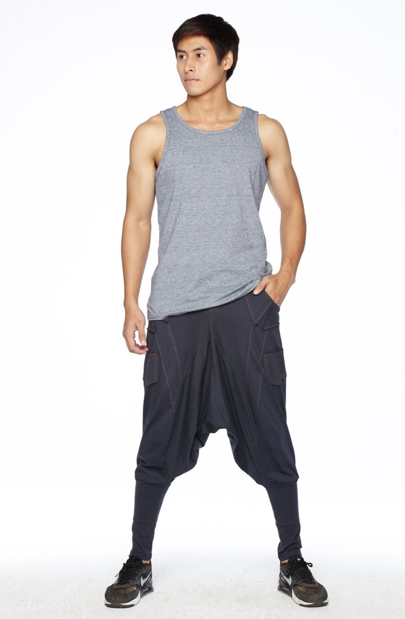 May include: A man wearing a gray tank top and dark blue pants with a unique design. He is standing with his hands in his pockets and looking to the side.