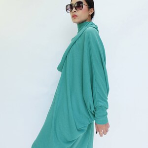 K&ouml;nnte beinhalten: Ein teal-farbenes, lang&auml;rmliges Kleid mit einem drapierten Rollkragen und einer lockeren, flie&szlig;enden Silhouette. Das Kleid wird mit einer gro&szlig;en, dunkel gerahmten Sonnenbrille kombiniert. Das Model steht vor einem wei&szlig;en Hintergrund.