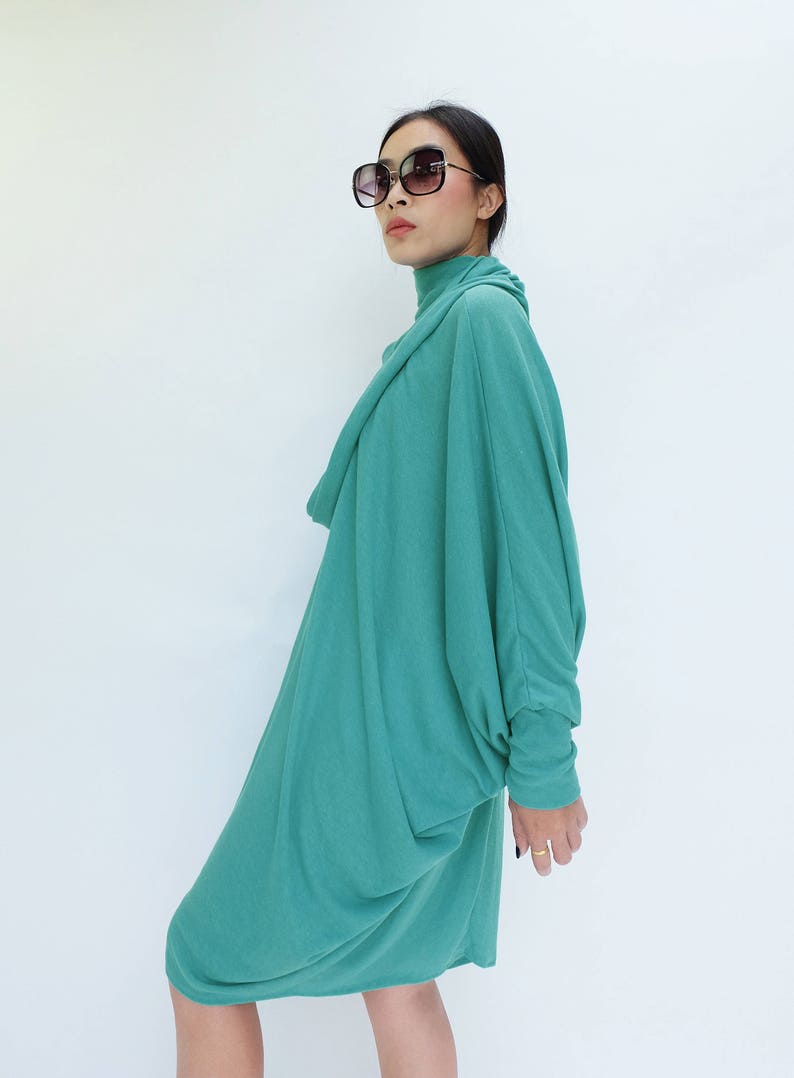 K&ouml;nnte beinhalten: Ein teal-farbenes, lang&auml;rmliges Kleid mit einem drapierten Rollkragen und einer lockeren, flie&szlig;enden Silhouette. Das Kleid wird mit einer gro&szlig;en, dunkel gerahmten Sonnenbrille kombiniert. Das Model steht vor einem wei&szlig;en Hintergrund.