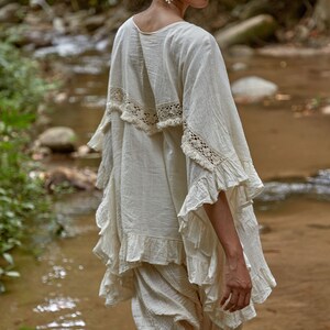 May include: A woman wearing a white cotton top with lace trim and ruffled sleeves. The top is paired with white cotton pants. The woman is standing in a stream.