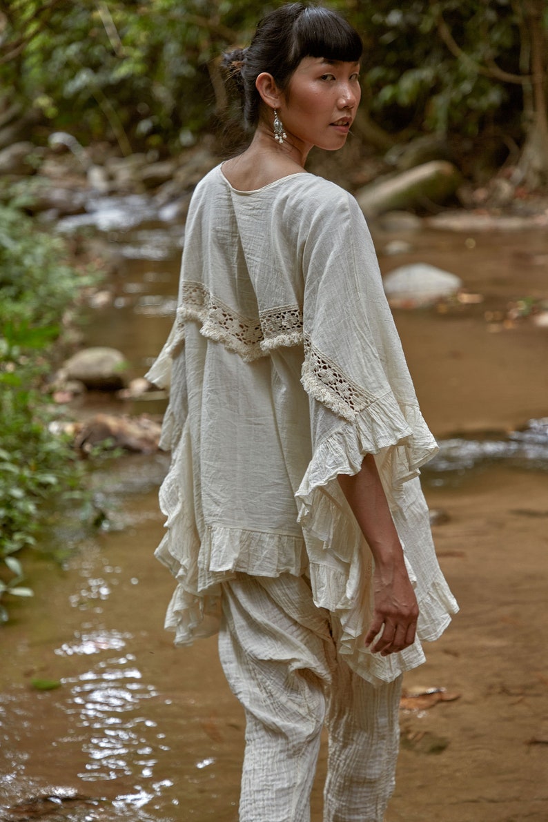May include: A woman wearing a white cotton top with lace trim and ruffled sleeves. The top is paired with white cotton pants. The woman is standing in a stream.